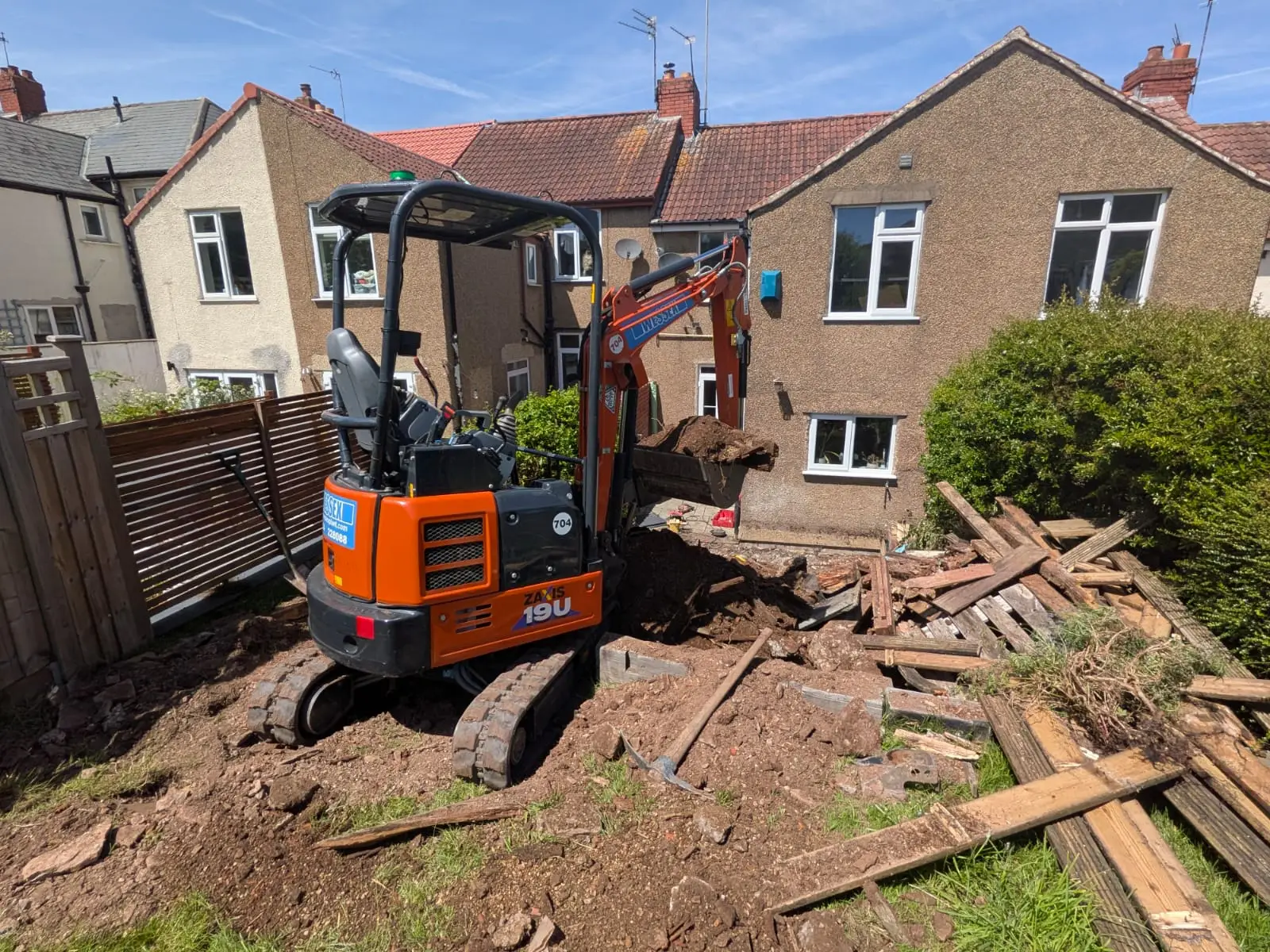 Mini digger stripping the ground of a rear garden, old timber stacked ready to go