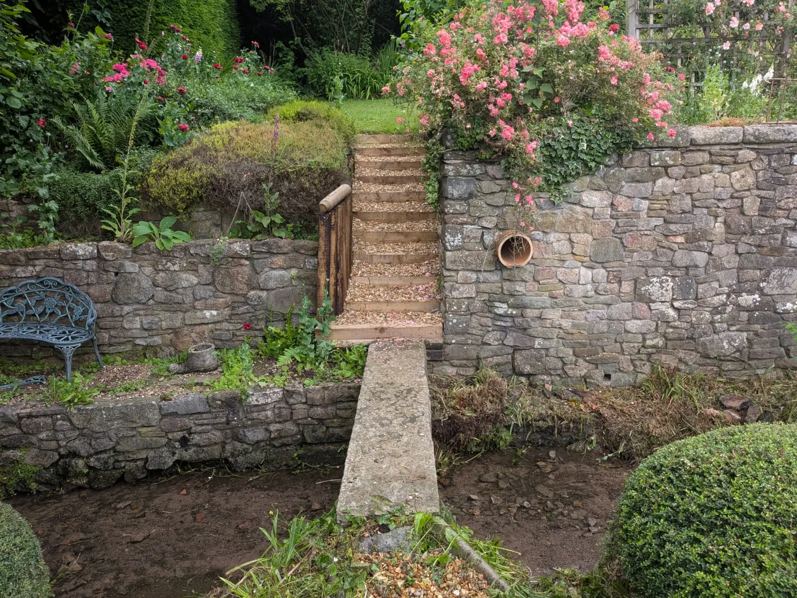 Dry stone retaining walls on two levels with timber steps and a stone slab bridge over a stream
